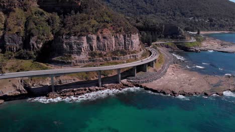 aerial view of cars driving on sea cliff bridge, sunny day, grand pacific drive, new south wales, australia - orbit drone shot