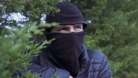 A-masked-thief-lurks-in-the-bushes,-revealing-an-automatic-weapon-from-his-dark-blue-jacket,-and-posing-a-potential-threat-to-a-individuals,-families-or-entire-neighborhoods