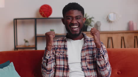 young adult man shouting, raising fists in gesture i did it, celebrating success, winning at home