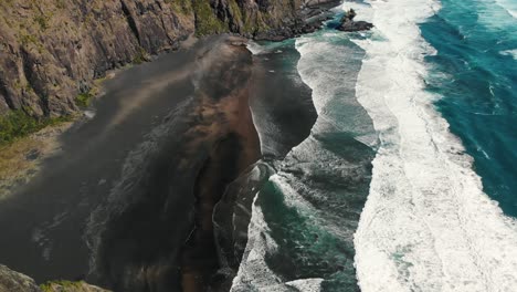 aerial-footage-waves-crash-on-the-beach-ocean-clear-water-and-rocky-cliff-in-a-natural-paradise,-new-zeland-Karekare