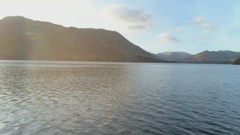 Low-Aerial-Drone-Shot-Flying-Over-Ullswater-Lake-at-Sunrise-on-Sunny-Morning-with-Hills-and-Mountains-in-Background-in-North-Lake-District-Cumbria-United-Kingdom