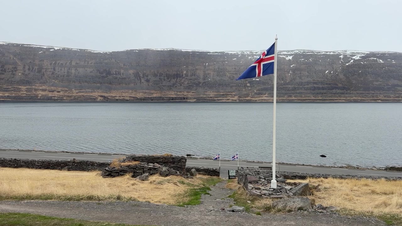 Et flagg på Island blafrer i vinden, plassert nær en fjord på Vestfjordene ved foten av noen små åser i et avsidesliggende område på Island
