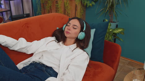 happy young woman in wireless headphones relaxing lying on sofa at home listening favorite music