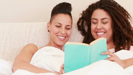una pareja de lesbianas sonrientes leyendo un libro en la cama.