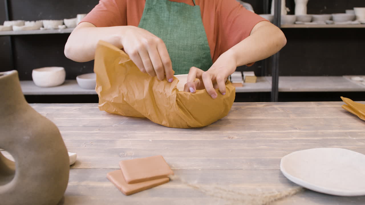 empleada irreconocible envolviendo cerámica artesanal con papel mientras se sienta a la mesa en la tienda de cerámica