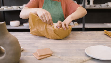 unerkennbare weibliche angestellte, die handwerkliche keramik mit papier umwickelt, während sie am tisch in der töpferei sitzt