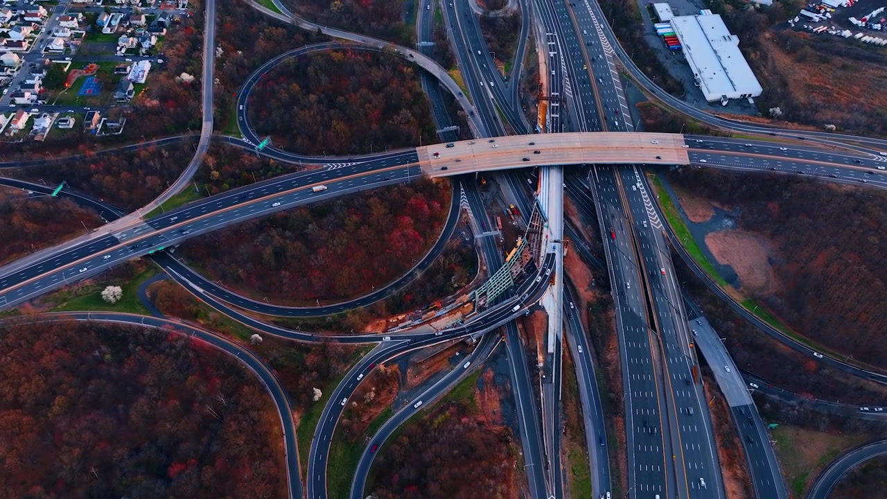 Top-down view of freeway junction beside a residential area, New Jersey. Overhead of an overpass and ramps weaving past autumn trees with houses and a warehouse along the edges