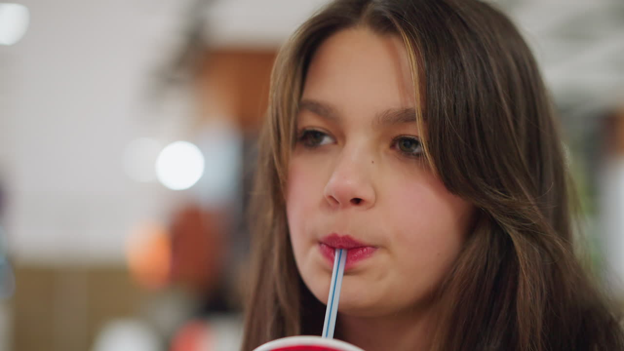 Close up of young woman with long brown hair holding drink straw as she interacts with someone off camera, showing natural candid expression against blurred indoor bokeh background