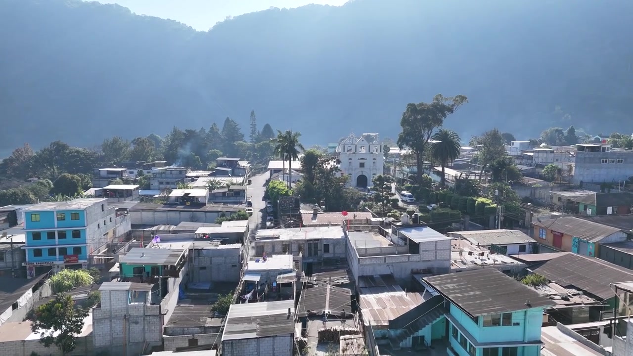 Vista aérea de drone sobre San Lucas, Guatemala, mostra bairros tropicais, palmeiras, céu claro, uma cordilheira vulcânica circundante e picos florestados formando um cenário natural deslumbrante para a cidade.