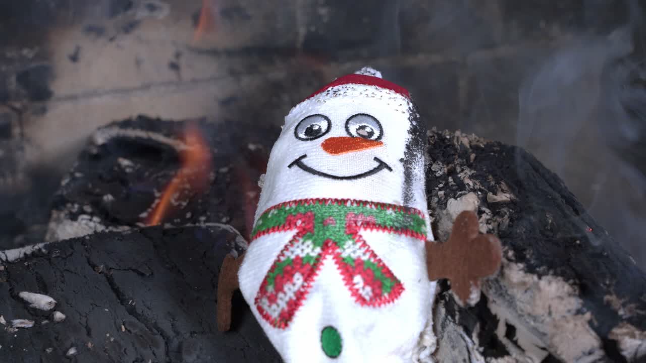 Cute small toy snowman IN red hat and green scarf from fabric put on burning coal in fireplace and burns macro