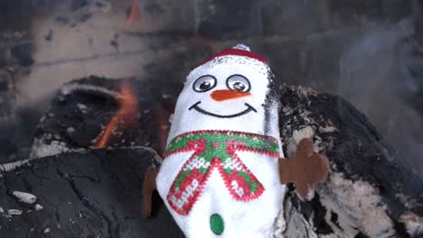 cute small toy snowman in red hat and green scarf from fabric put on burning coal in fireplace and burns macro