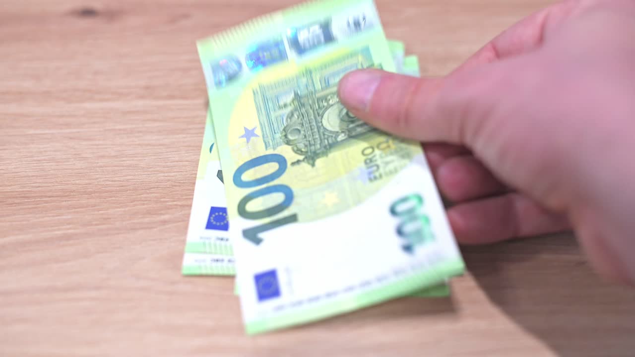 A close-up video of several €100 Euro banknotes spread across a wooden table, with a hand holding one of the bills in the foreground. The image emphasizes cash, wealth, and financial concepts.