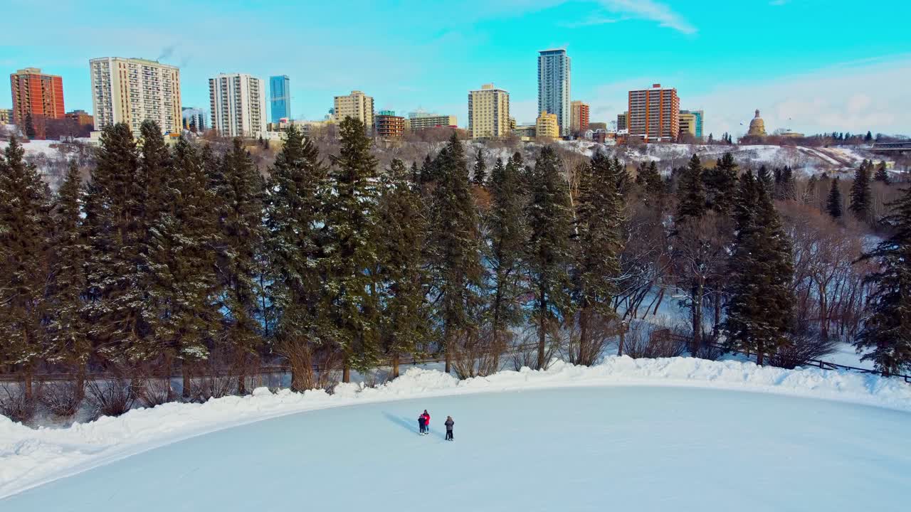 una pareja de citas dobles soporta el clima frío calentándose mutuamente en la pista de patinaje sobre hielo artificial del parque victoria en primer plano con apartamentos en condominio en el horizonte detrás de la nieve de un pino alto edm2-4