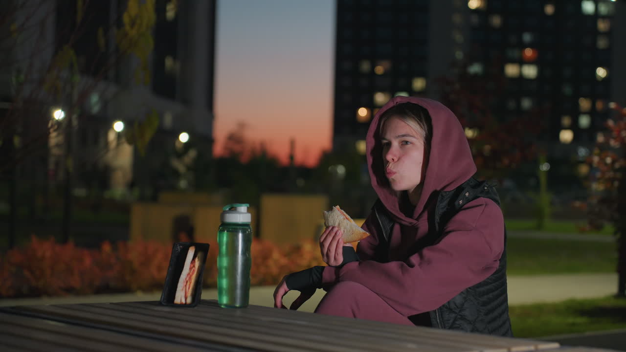 Fair skinned woman wearing hoodie sits on bench outdoors at dusk holding sandwich in hands under bright lighting next to water bottle on table while city lights glow in blurred urban background