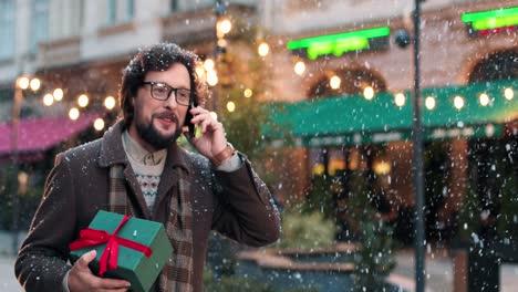 Close-up-view-of-caucasian-man-wearing-eyeglasses-holding-a-present-and-talking-on-smartphone-on-the-street-while-it‚Äôs-snowing-in-Christmas