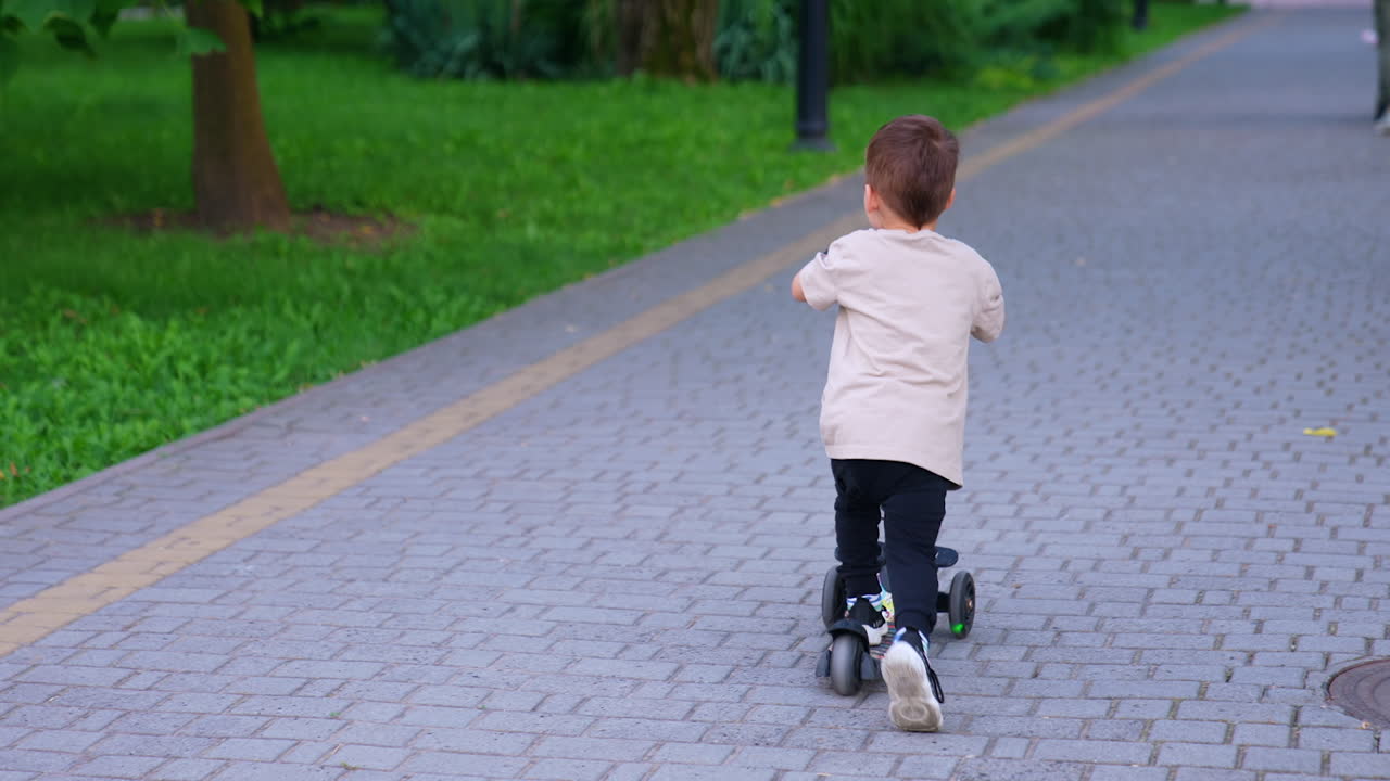 Rear view of a baby boy riding on the scooter. Kid points at the something in the park with a finger.