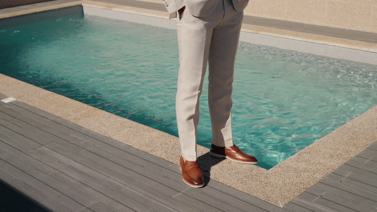 hombre con pantalones formales beige y zapatos marrones pulidos de pie junto a una lujosa piscina con agua azul clara
