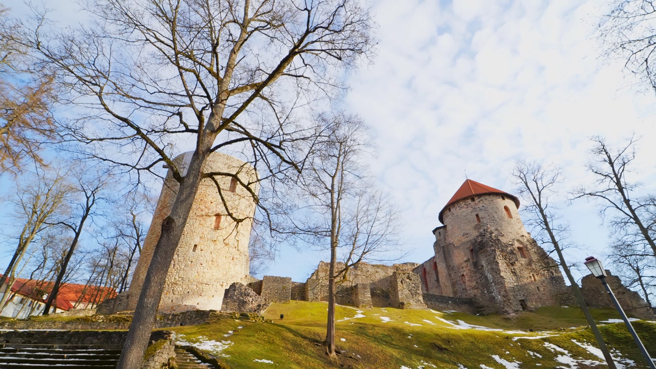 Cesis Castle towers and ruins above stone stairs and a small bridge in winter sunlight, with melting snow and bare trees in a scenic historic landscape.