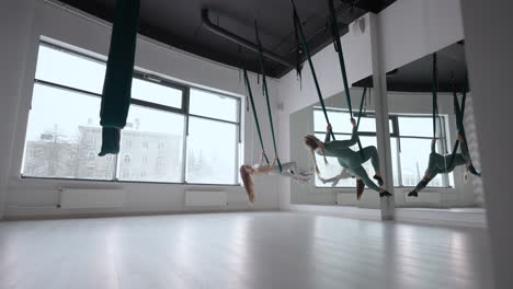 trainer and young woman practising aero gymnastics with suspensions at gym. group of two young beautiful yogi women doing aerial yoga practice in green hammocks in fitness club