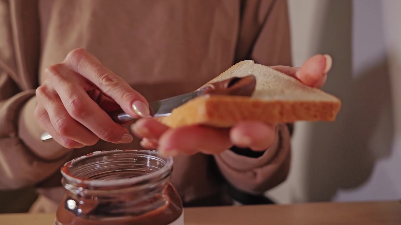 Woman picks up a slice of white bread and begins spreading sweet chocolate paste