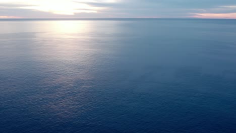 an endless blue ocean stretching out to the horizon at dusk - aerial pull back flyover