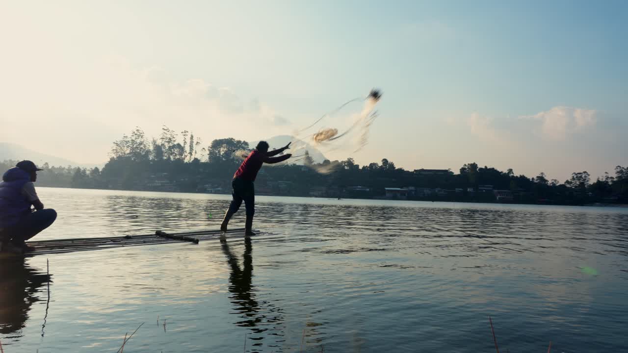Two Fishermen On Bamboo Raft Throwing A Fishing Net Into The Calm Lake Under A Beautiful Sunrise And Blue Sky With Scattered Clouds