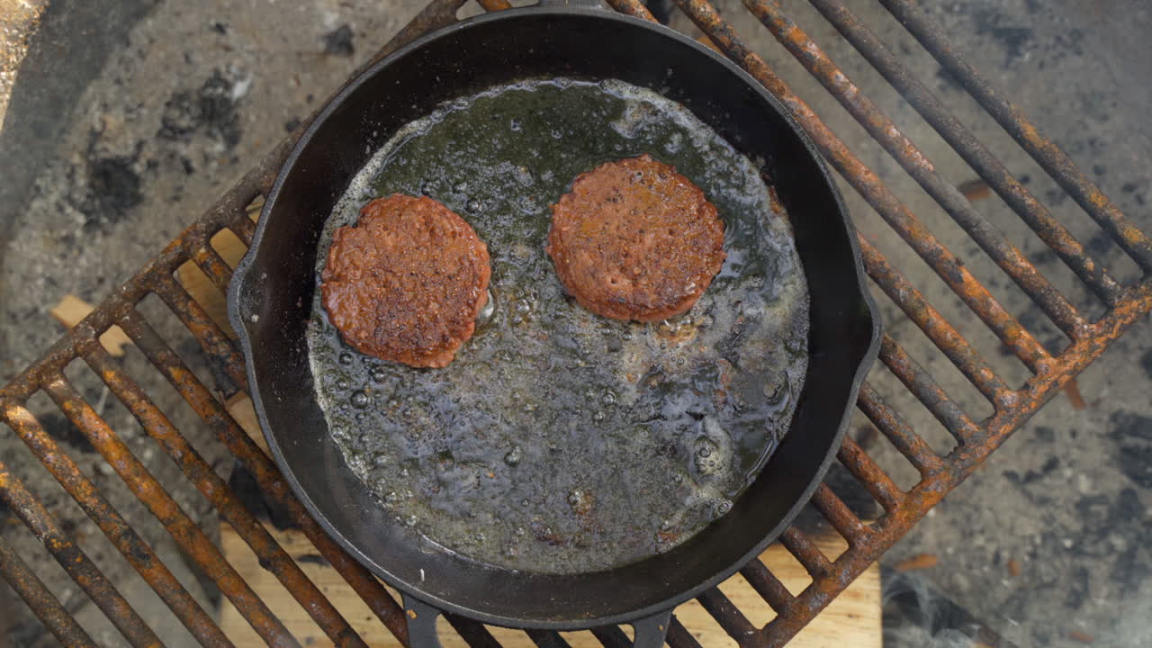 캠핑 여행 중 숲 한가운데 연기가 자욱한 모닥불 위에 뜨겁고 검은 철 프라이팬에서 요리하는 야채 버거의 오버헤드 뷰를 닫습니다.