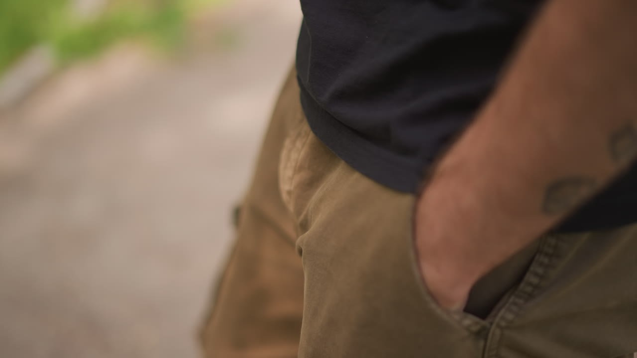 White Man Showing Hand Gesture Casually While Pocketing Fingers, Close View Of Khaki Pants And Tattooed Forearm, Relaxed Street Style Posture In Park Setting, Subtle Attitude And Body Language