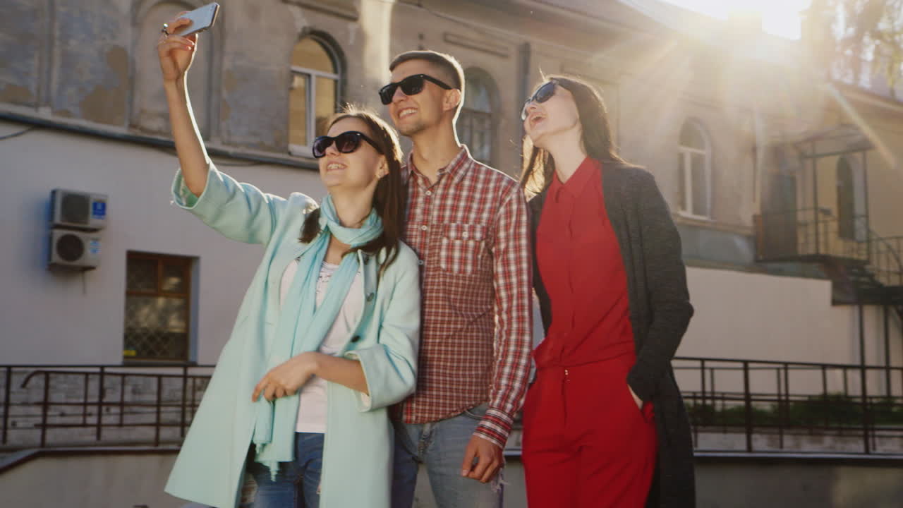 amigos dos mujeres y un hombre haciendo selfie bajo el sol contra el fondo de un viejo edificio hd vid