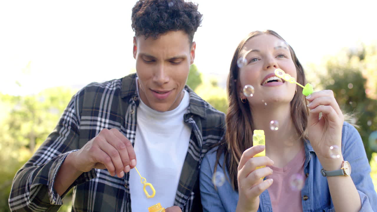 una pareja feliz y diversa soplando burbujas en un jardín soleado, en cámara lenta.
