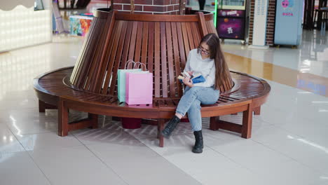 woman seated on a circular wooden bench in a well-lit mall, leg crossed, deeply engrossed in a book, with green light shining in the background and shopping bags beside her