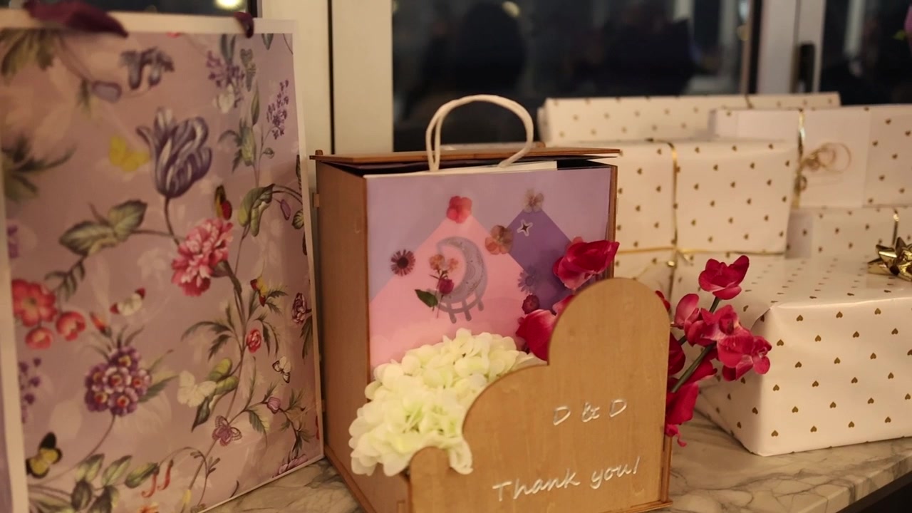 Primo piano mostra sacchetti regalo splendidamente disposti, fiori delicati e confezioni eleganti su una superficie di marmo, illuminati calorosamente in un ambiente interno serale, evocando celebrazione e gratitudine