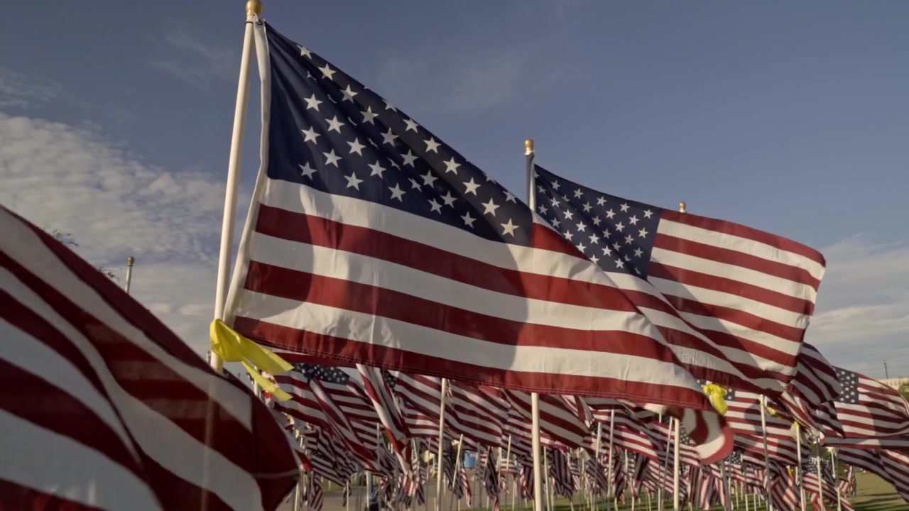 Rows of American flags wave heavily in the wind at an outdoor memorial park near sunset. The static shot captures the patriotic display against a sky conveying honor and remembrance.