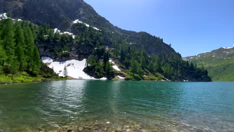 Toma-Panorámica-Del-Lago-Natural-Entre-Montañas-Austriacas-Cubiertas-De-Nieve-En-Verano