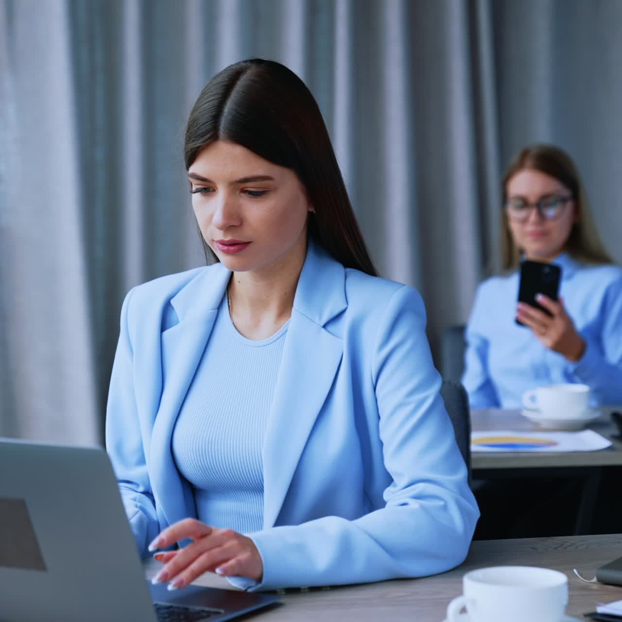 Busy employees working at office sitting at desks in front of computers. Lady at desk behind looks at her phone. Blurred backdrop