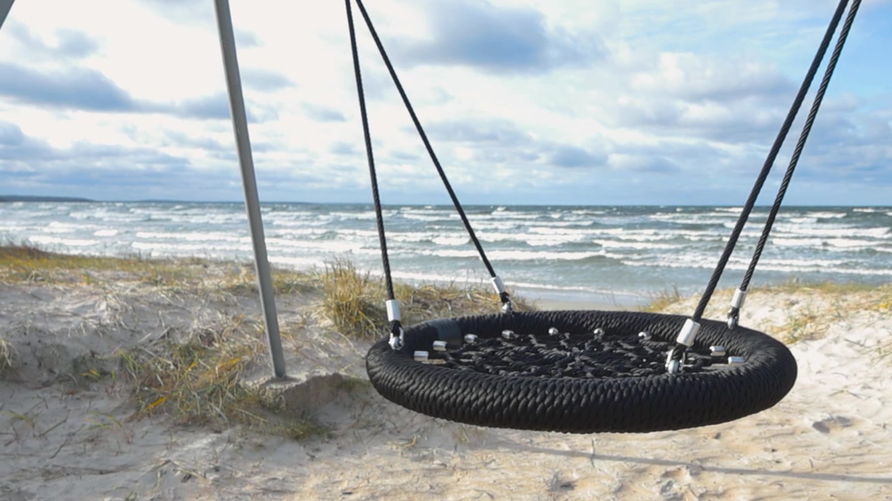 Swing at a beach swinging and rocking in the wind while Baltic ocean sea water waves are splashing and moving in the background in slow motion during a cloudy day with dark clouds in the sky. Windy.