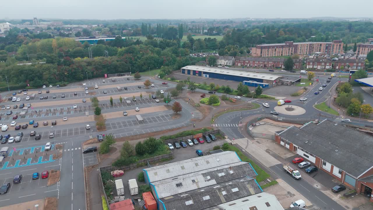 Aerial view of an urban area with parking lots and industrial buildings under an overcast sky. The scene includes cars, roads, and trees, showcasing the blend of infrastructure and natural elements.
