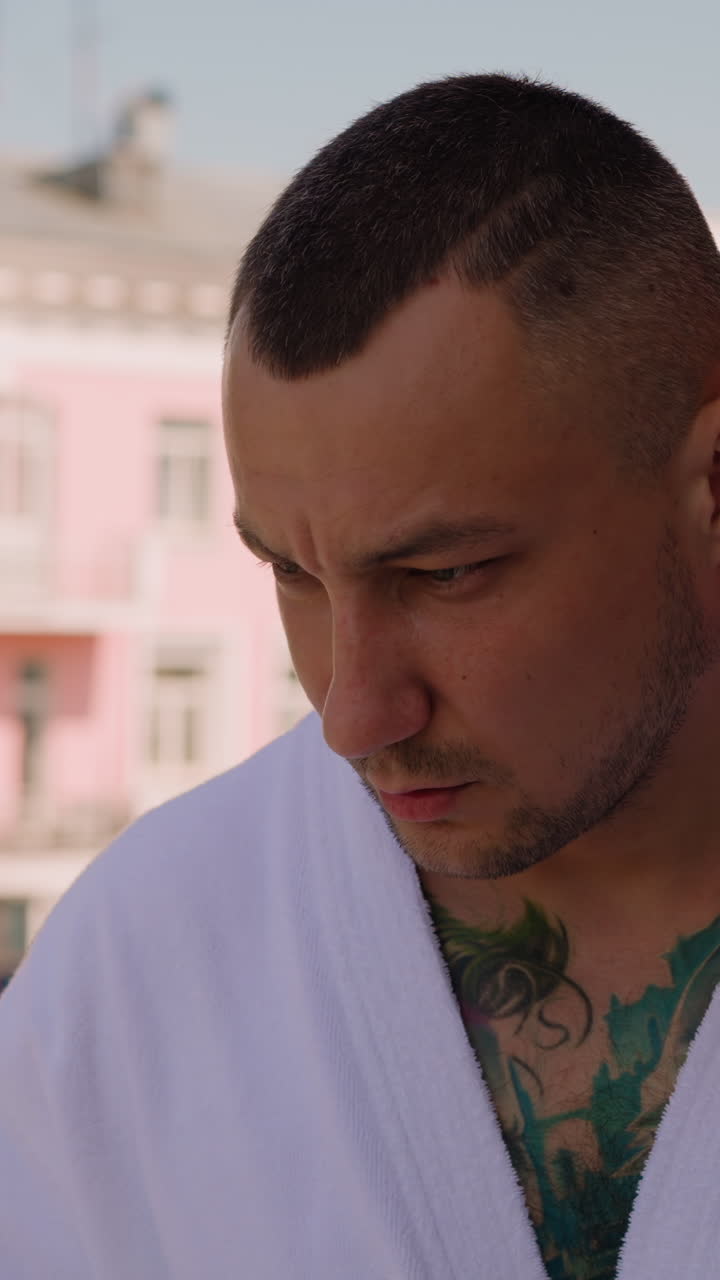 Upset man with colorful tattoos on body wearing white bathrobe stands looking down on balcony of hotel in large city closeup slow motion