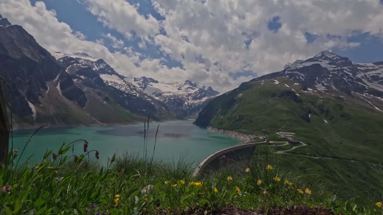 Timelapse of a stunning mountain reservoir is surrounded by towering snow-capped peaks and vibrant wildflowers in the foreground. A curved dam stretches across the turquoise water.