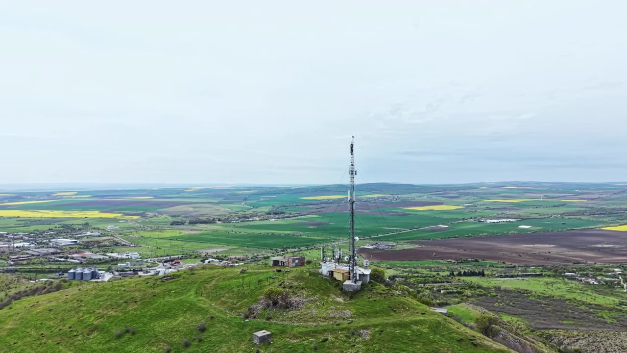Utsikt over et kommunikasjonstårn på en ås i et landlig område med jorder og en skyet himmel i bakgrunnen.