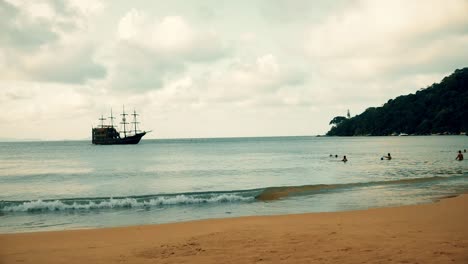 Un-Barco-Medieval-Se-Acerca-A-La-Playa-Para-Atracar-En-El-Puerto-En-Un-Día-Nublado-Con-Gente-En-El-Agua