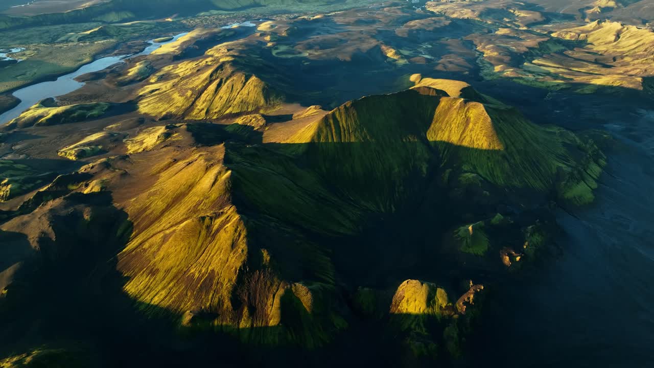 Amplio sobrevuelo de dron por las tierras altas de Islandia a la hora dorada, colinas volcánicas verdes iluminadas por el sol y planicies de lava oscuras con lagos serpenteantes, sombras largas, cielo despejado y en calma, serenidad épica