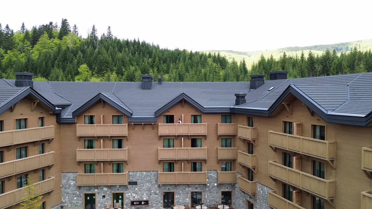 Woman standing on balcony of modern wooden chalet surrounded by lush pine forest in summer, Biogradska Gora National Park, Aerial revealing shot