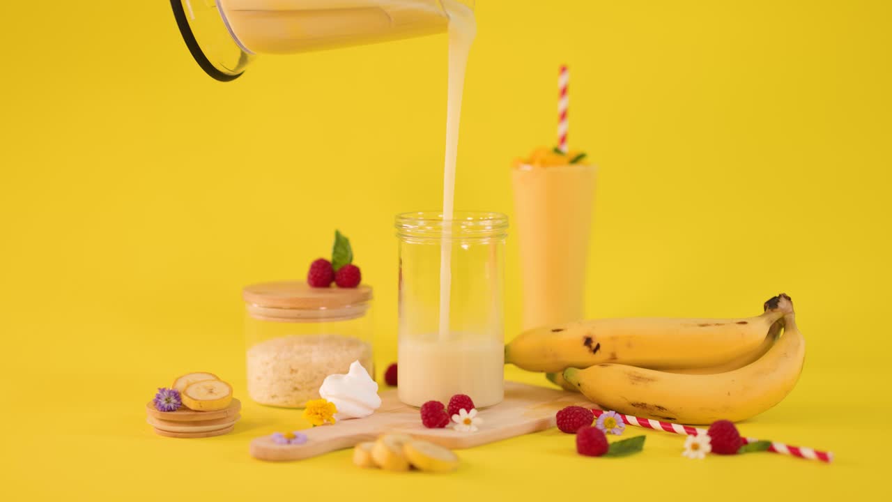 Smoothie cremoso alla banana versato in un bicchiere tra frutta fresca e dessert su sfondo giallo brillante