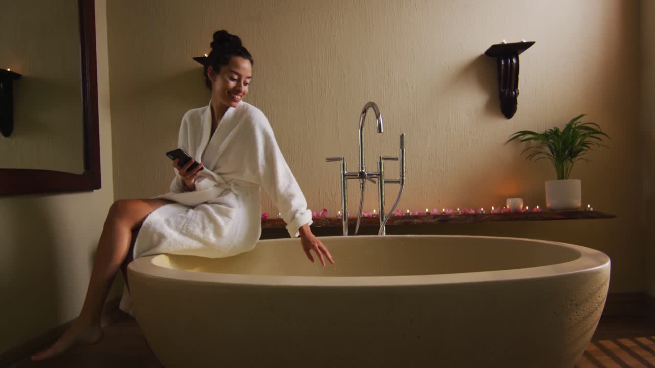 счастливая двухрасовая женщина сидит в халате на ванной, готовит ванну, использует смартфон