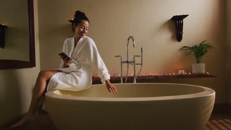 happy biracial woman sitting in robe on bathtube, preparing bath, using smartphone