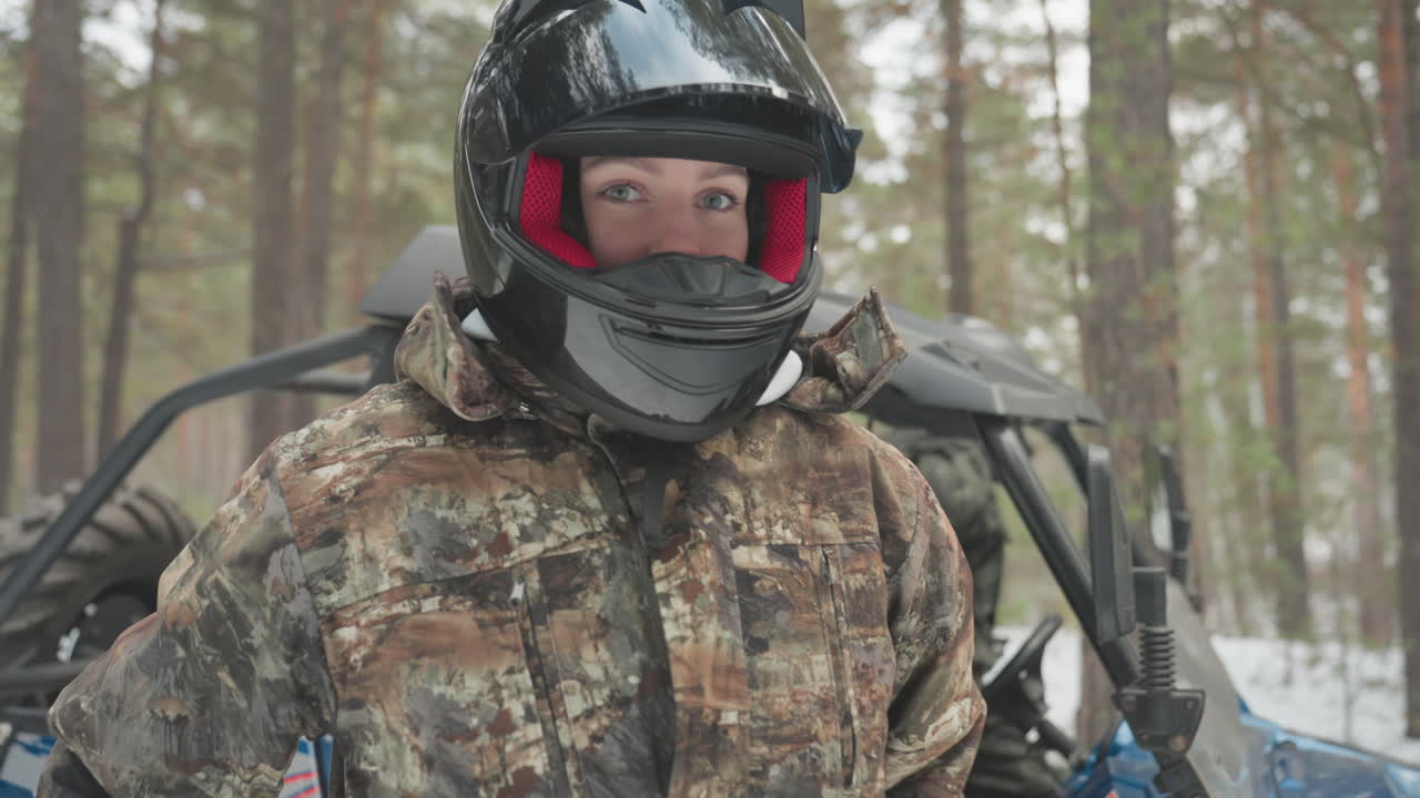 Pilota mimetizzato accanto all'ATV in una foresta innevata che si prepara per una corsa fuoristrada estrema, indossa un casco nero con occhiali rossi e una giacca mimetica coperta di neve, regolando lo sterzo e l'equipaggiamento di sicurezza