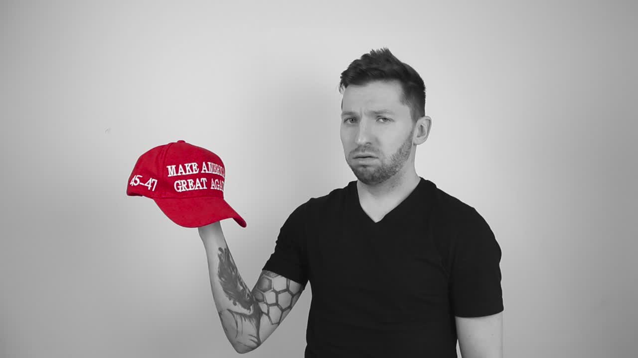 Grayscale footage of a man with tattoos, short hair and a beard lifting up a red vibrant colored MAGA Make America Great Again hat and blowing air out of his mouth indicating he really hates it.