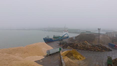 aerial establishing view of wood terminal crane loading timber into the cargo ship, port of liepaja , lumber log export, day with fog and mist, wood chip piles, wide drone shot moving forward