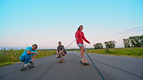 Front-View-Of-Two-Boys-And-A-Girl-Skateboarding-Holding-On-To-A-Rope-On-The-Road,-In-The-Sky-A-Parachutist-Flying